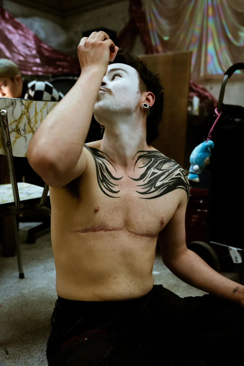 Performer met witte schmink en een borsttatoeage die backstage omhoog kijkt