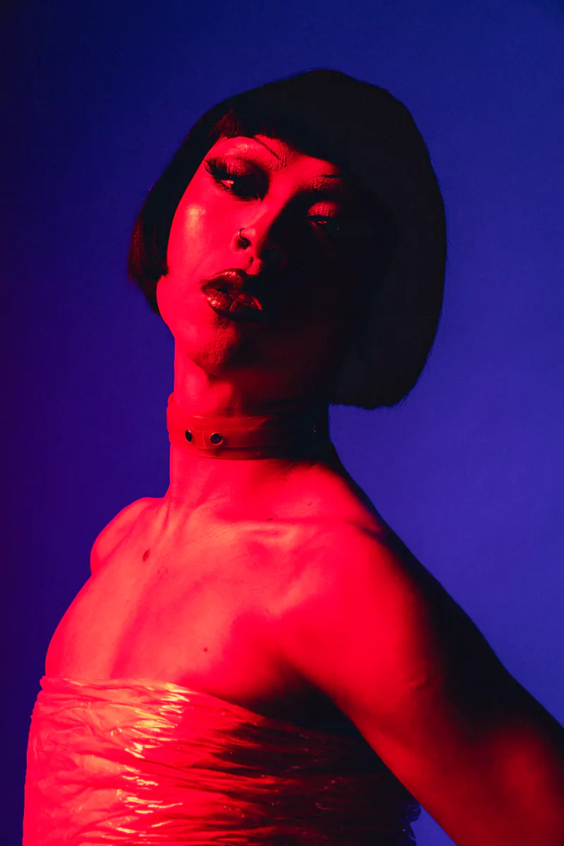 Portret in rood en blauw licht, lichaam gewikkeld in plasticfolie, met bobpruik en halsband